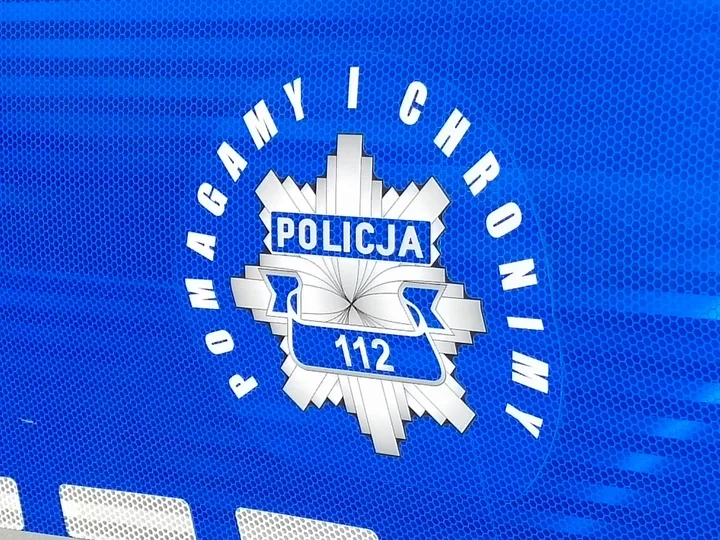 Fragment niebieskiego pojazdu Policji z białym emblematem z napisem „POLICJA”, numerem alarmowym 112 i hasłem „POMAGA MY I CHRONIMY”.