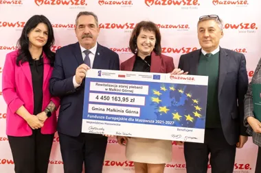 Cztery osoby stoją przed ścianką z logo Mazowsze i trzymają duży czek na dofinansowanie rewitalizacji starej plebanii w Małkini Górnej z funduszy europejskich.